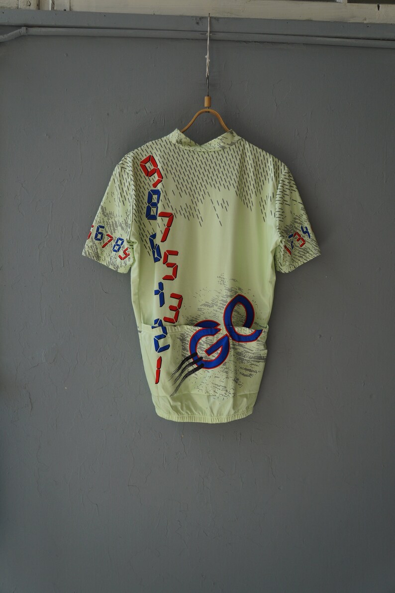 May include: A light green cycling jersey with a blue and red graphic design. The design features a number sequence and the letter "G" repeated twice.