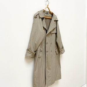 May include: A long, beige trench coat with a double-breasted closure and a hood. The coat is hanging on a wooden hanger against a white wall.