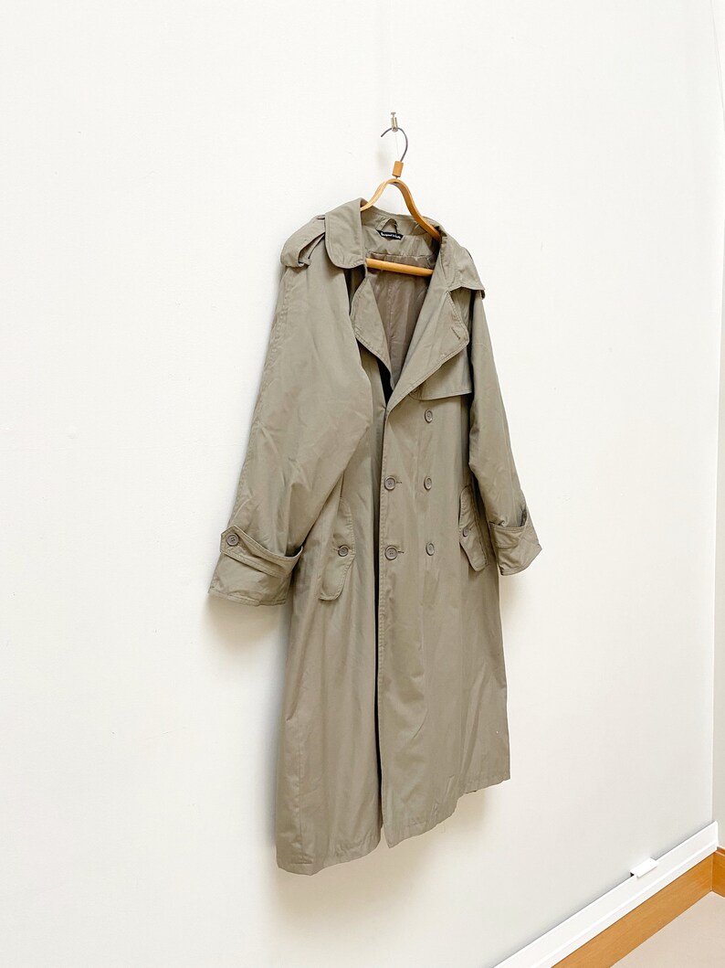 May include: A long, beige trench coat with a double-breasted closure and a hood. The coat is hanging on a wooden hanger against a white wall.