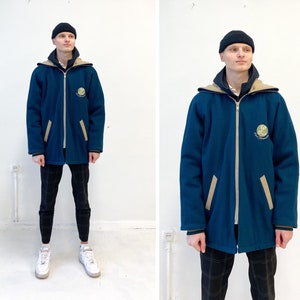 Op de afbeelding: Een blauwe jas met een beige kraag en beige accenten. De jas heeft een ritssluiting en een logo op de linkerborst. De persoon draagt een zwart-wit geblokte broek en witte sneakers.