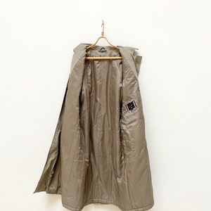 May include: A long, khaki-colored trench coat with a button closure and a small patch on the right side. The coat is hanging on a wooden hanger.