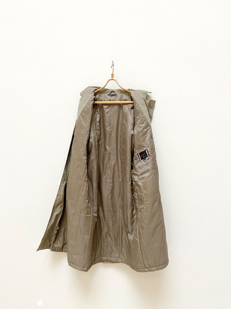 May include: A long, khaki-colored trench coat with a button closure and a small patch on the right side. The coat is hanging on a wooden hanger.