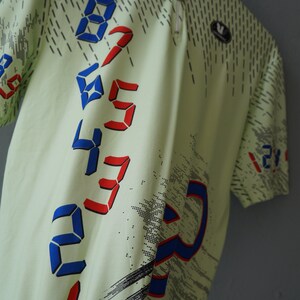 May include: A light green short-sleeved shirt with a digital print design. The shirt features a pattern of red and blue numbers in a diagonal line. The numbers are 0, 1, 2, 3, 4, 5, 6, 7, 8, and 9.