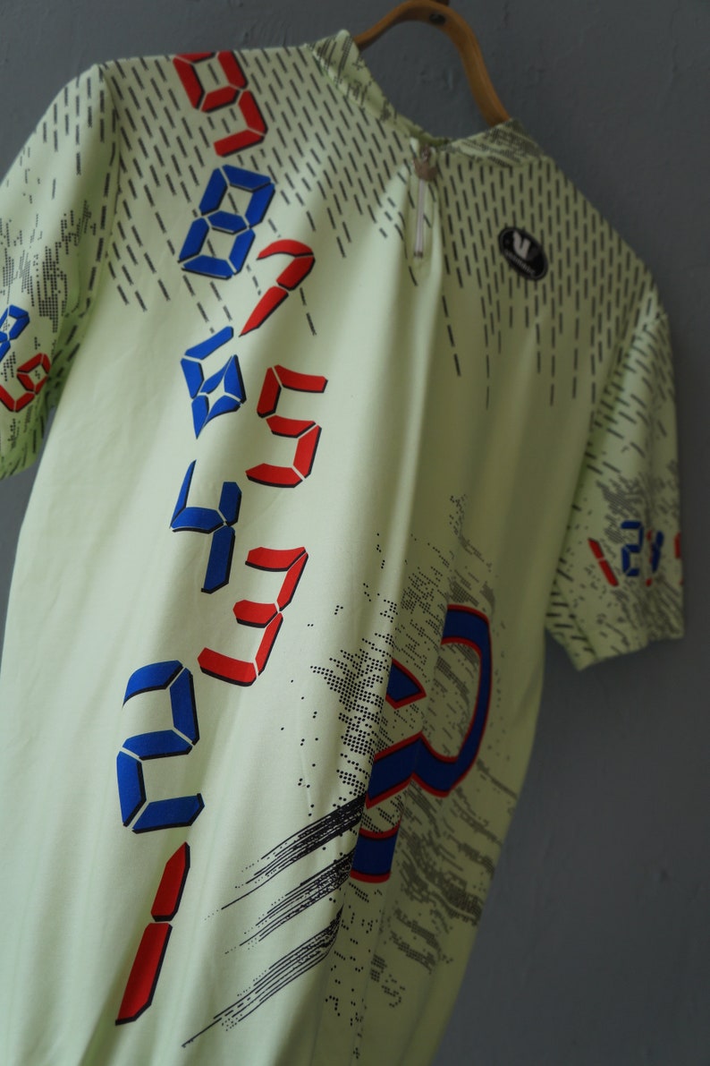 May include: A light green short-sleeved shirt with a digital print design. The shirt features a pattern of red and blue numbers in a diagonal line. The numbers are 0, 1, 2, 3, 4, 5, 6, 7, 8, and 9.