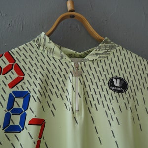May include: A light green zippered shirt with a repeating pattern of the word "VERMARC" in black. The shirt has a digital clock design with red and blue numbers.