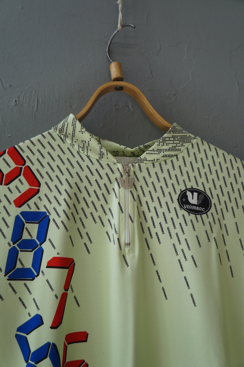 May include: A light green zippered shirt with a repeating pattern of the word "VERMARC" in black. The shirt has a digital clock design with red and blue numbers.