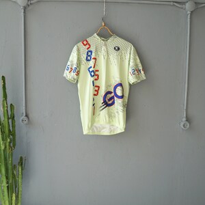 May include: A light green cycling jersey with a white, blue, and red design. The jersey has a zipper closure and the word "GO" printed on the front. The numbers 1 through 9 are printed on the left side of the jersey and the numbers 1 through 4 are printed on the right side.