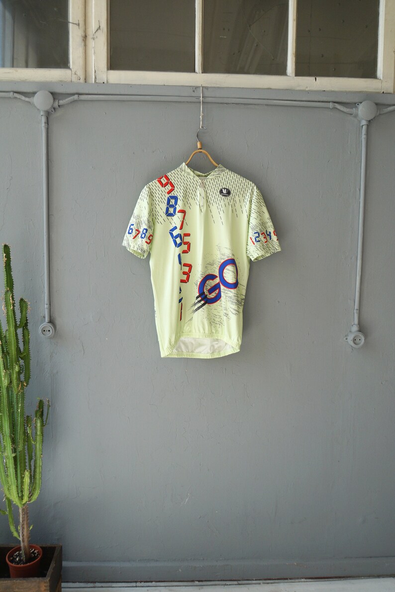 May include: A light green cycling jersey with a white, blue, and red design. The jersey has a zipper closure and the word "GO" printed on the front. The numbers 1 through 9 are printed on the left side of the jersey and the numbers 1 through 4 are printed on the right side.
