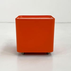 Könnte beinhalten: Eine leuchtend orangefarbene, quadratische Aufbewahrungseinheit mit abgerundeten Ecken und einer flachen Oberseite. Das Möbelstück hat eine glatte, einfarbige Oberfläche und steht auf kleinen, unauffälligen Rollen an der Unterseite.