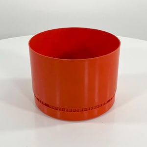 May include: A cylindrical, orange container with a textured band near the base. The interior is a vibrant red. The container is made of a smooth, solid material and sits on a white surface. The design is simple and modern.