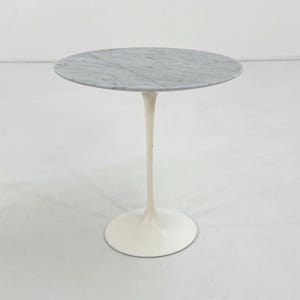May include: A round side table featuring a grey and white marble top with natural veining. The table is supported by a single, slender, off-white pedestal base that flares out at the bottom into a circular floor plate, creating a stable structure.