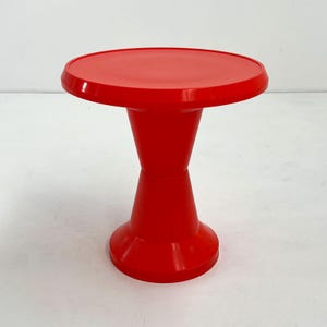 May include: A bright red plastic pedestal stand featuring a smooth, circular top surface and a central support column that tapers inward toward the middle, resting on a wide, stable circular base. The entire structure is a solid, uniform red color.