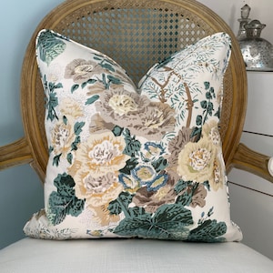 May include: A decorative throw pillow with a floral pattern featuring large cream, tan, and blue flowers with green leaves. The pillow is on a light-colored chair with a woven back. The pillow has a white background.