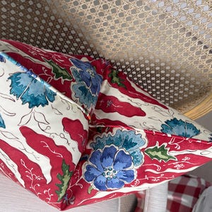 May include: Two decorative throw pillows with a floral pattern. The pillows feature a white background with red stripes and blue floral designs. The pillows are placed on a light-colored chair with a woven back.