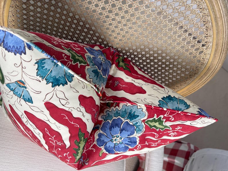 May include: Two decorative throw pillows with a floral pattern. The pillows feature a white background with red stripes and blue floral designs. The pillows are placed on a light-colored chair with a woven back.