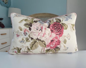 May include: Rectangular cushion with a floral design. The cream-coloured fabric is patterned with large pink and red roses, green leaves, and smaller blue and white flowers. The edges are trimmed with pink piping.