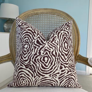 May include: A decorative throw pillow with a white background and a repeating pattern of dark brown concentric circles. The pillow is placed on a white upholstered chair with a light brown wooden frame and a woven back.