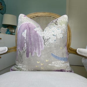 May include: Decorative pillow with a landscape design featuring a purple weeping willow tree, white flowering trees, and a blue boat on a gray background. The pillow is placed on a white chair with a gold-colored frame.