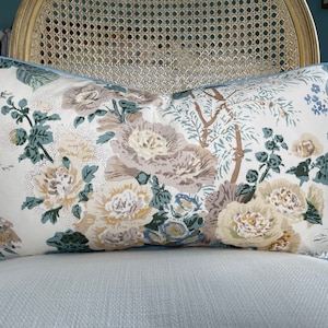 May include: Rectangular decorative pillow with a floral pattern in cream, beige, and blue hues. The pillow features large, stylized flowers and foliage, with a light blue border. The pillow is placed on a white chair.