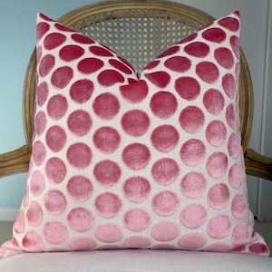 May include: A decorative throw pillow with a white background and a pattern of large, pink polka dots. The pillow is placed on a white chair with a light brown, ornate wooden back.