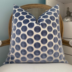 May include: A square decorative pillow with a cream-colored background and large, evenly spaced blue polka dots. The pillow is placed on a vintage-style chair with a light wood frame and a white seat.