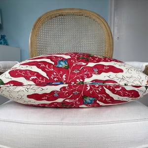 May include: A decorative pillow with a red and white floral pattern, featuring red wavy stripes and blue floral accents. The pillow is placed on a white cushion and a wooden chair with a woven back.
