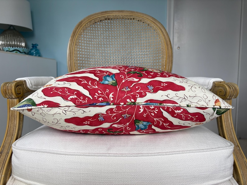 May include: A decorative pillow with a red and white floral pattern, featuring red wavy stripes and blue floral accents. The pillow is placed on a white cushion and a wooden chair with a woven back.