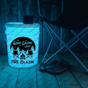May include: A glowing white bucket with the text "Star Gazin' and Fire Blazin'" in black. The bucket has a black graphic of a campfire, chairs, and trees. A black mug sits on top. A folding chair and sandals are in the background.