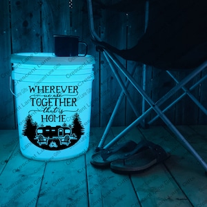 May include: A white bucket illuminated with a soft glow, featuring the text "WHEREVER we are TOGETHER that is HOME" and a camping design. A black mug rests on top. A folding chair and sandals are nearby.