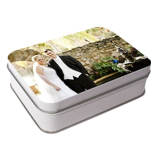 May include: A white metal box with a photo of a bride and groom on the lid. The photo shows the couple standing in front of a stone wall with a fountain in the background.