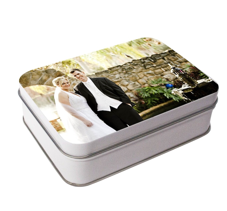 May include: A white metal box with a photo of a bride and groom on the lid. The photo shows the couple standing in front of a stone wall with a fountain in the background.