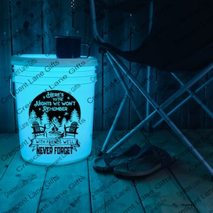 May include: A white bucket illuminated with a blue glow, featuring the text "Here's to the nights we won't remember, with friends we'll never forget." A black mug sits on top. A folding chair and sandals are nearby.