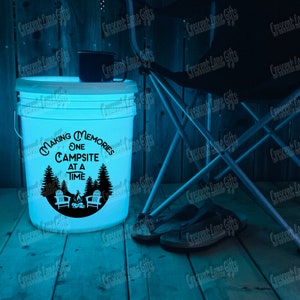 May include: A glowing white bucket with the text "Making Memories One Campsite at a Time" and a camping scene graphic. A black mug sits on top. A folding chair and sandals are nearby, suggesting a camping theme.