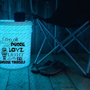 May include: A white bucket with the text "I'm all peace, love, light and a little go smudge yourself" in black. A black mug sits on top of the bucket. A folding chair and sandals are in the background.