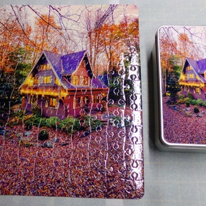 May include: A completed jigsaw puzzle of a house with lights in the woods. The puzzle is in a metal tin with the same image on the lid.