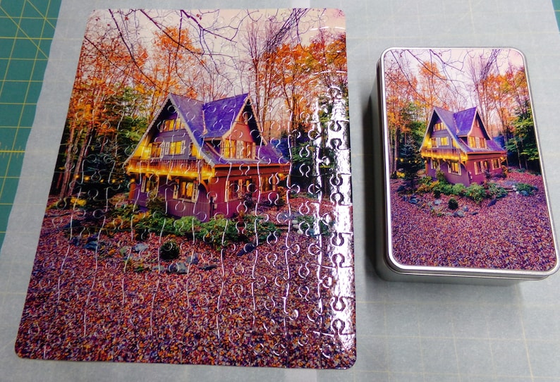 May include: A completed jigsaw puzzle of a house with lights in the woods. The puzzle is in a metal tin with the same image on the lid.