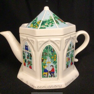 May include: White ceramic teapot shaped like a conservatory with a blue and green roof and colourful floral designs. The teapot has a handle and spout and reads "a conservatory teapot".