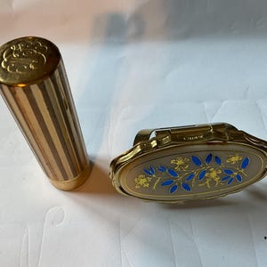 May include: A gold compact with a floral design and blue accents. The compact is open and the inside is visible. There is also a gold lipstick tube with a striped pattern.