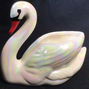 May include: A white iridescent ceramic swan planter with a red beak and black eyes. The swan's neck is curved and the body is hollowed out to form a planter.