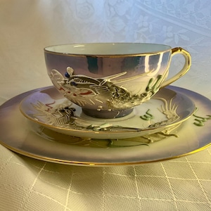 May include: A porcelain tea set featuring a dragon design. The set comprises a teacup, saucer, and a small plate, all with a pearlescent sheen. The dragon is sculpted in relief, with gold trim on the cup handle and edges.