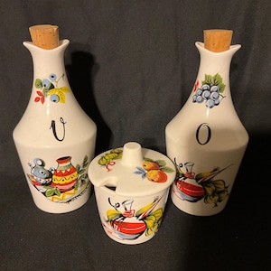 May include: A set of three vintage ceramic condiment bottles with cork stoppers. The bottles are white with hand-painted floral and fruit designs. The bottles are labelled "V" and "O".