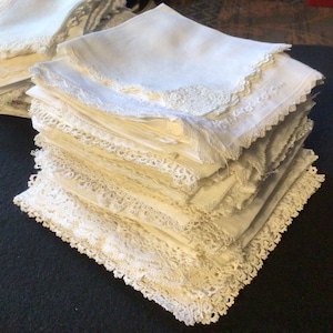 May include: A stack of white linen handkerchiefs with lace trim. The handkerchiefs are folded and stacked neatly, with the lace trim visible on the edges.