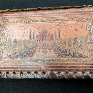 May include: A brown leather wallet with a decorative design of a building with trees and a sky. The wallet has a braided leather border.