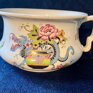 May include: A white ceramic chamber pot featuring a colourful transferware design of a blue dragon, a green and black patterned vessel, and pink and yellow floral motifs. The vessel has a wide rim, a curved body, and a single side handle.