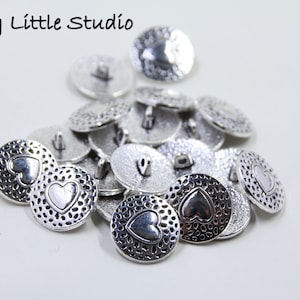 May include: A pile of silver buttons, each featuring a heart motif. The buttons have a textured surface and a loop on the reverse for sewing. The text "My Little Studio" is visible in the top left corner.