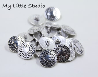 May include: A collection of silver-colored buttons with a heart design. The buttons have a textured, hammered appearance and a small loop on the back for sewing. The text "My Little Studio" is in the upper left corner.