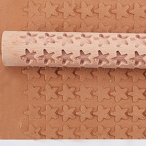 Puede incluir: Se muestra un rodillo de madera con cortes en forma de estrella, presionado sobre una superficie marrón, creando un patrón de estrellas repetitivo. El rodillo es de color marrón claro y cilíndrico, diseñado para imprimir diseños en masa. La superficie es de un marrón terroso sólido.