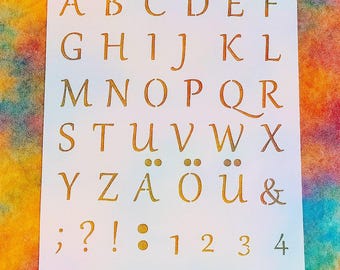 May include: A white stencil with the alphabet, numbers, and symbols in a gold color. The stencil includes the letters A through Z, the numbers 0 through 9, and punctuation marks. The background is a colorful, abstract design.