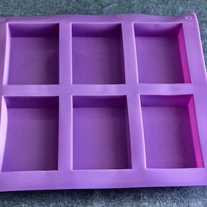 Puede incluir: Un molde cuadrado de silicona morada con seis cavidades rectangulares. El molde tiene bordes redondeados y pequeños agujeros circulares en cada esquina. Este artículo se utiliza probablemente para hacer jabones, velas u otras manualidades.