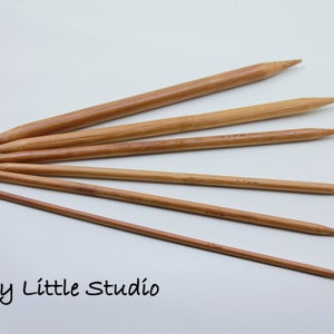 Könnte beinhalten: Sechs Stricknadeln aus Holz in verschiedenen Größen auf einer weißen Oberfläche. Die Nadeln sind hellbraun und haben spitze Enden. Einige Nadeln haben Größenangaben in mm.